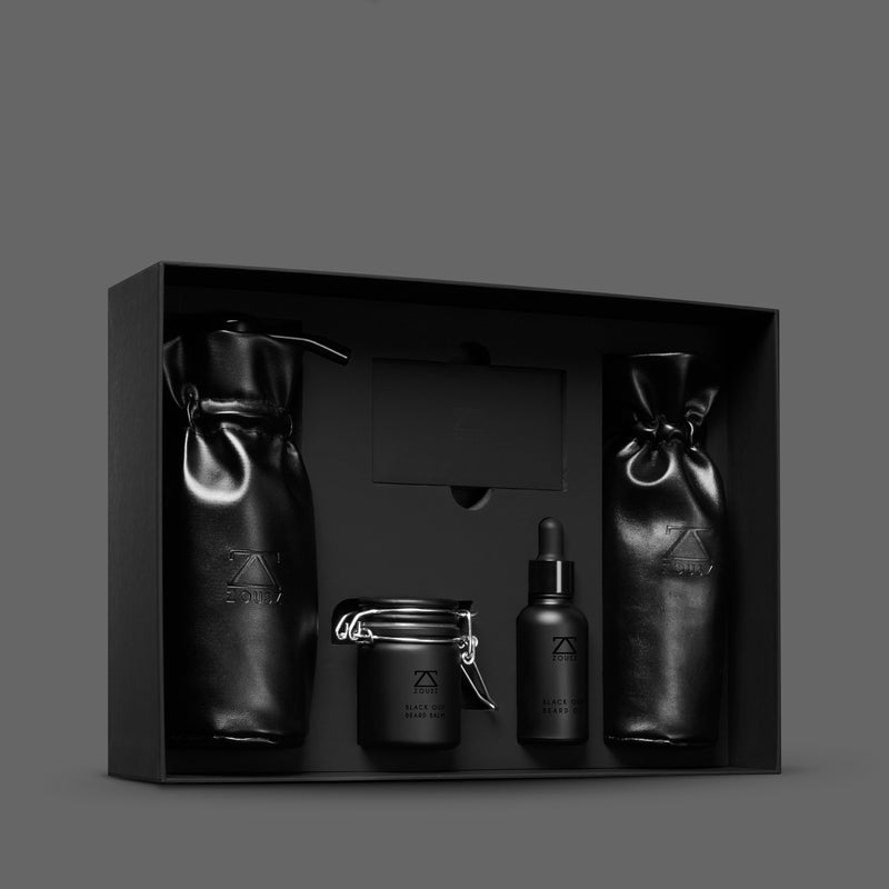 The black oud collection. A black skincare gift set displayed inside a black presentation box, featuring two matte black drawstring pouches, a small black dropper bottle, and a black jar with a metal clasp lid, all on a dark grey background.